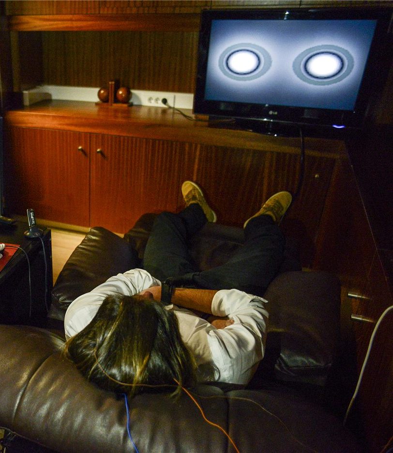 A person reclined on a sofa wearing an EEG headset, facing a screen with a feedback display