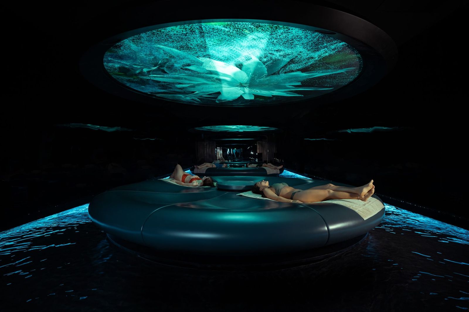 Two people reclining in a circular wellness pod under a teal ceiling