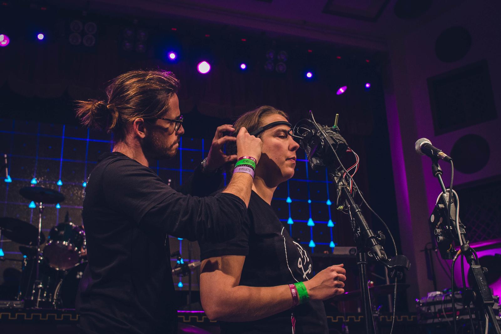 On a lit stage, one performer adjusts an EEG headset on another