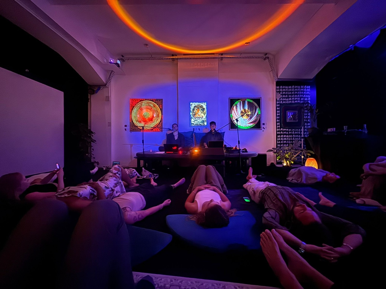 A group lying on cushions in a violet-lit room under an orange light ring during a breath session